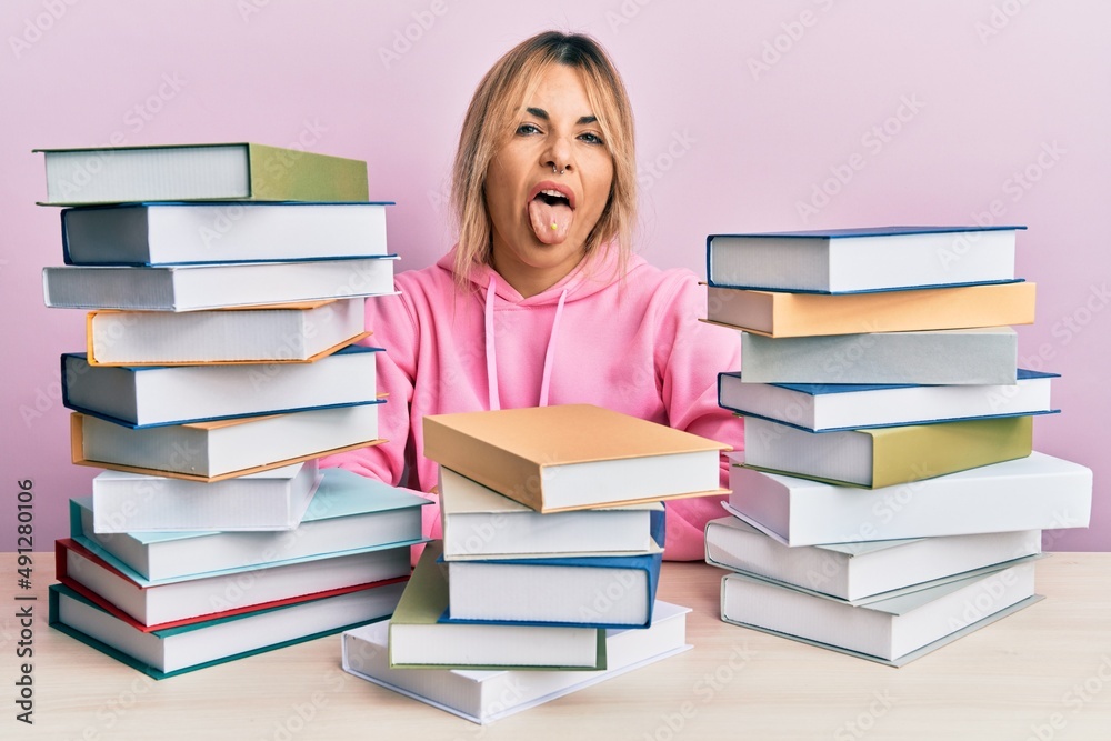 Young caucasian woman sitting on the table with books sticking tongue out happy with funny expression. emotion concept. Young caucasian woman sitting on the table with books sticking tongue out happy with funny expression. emotion concept.