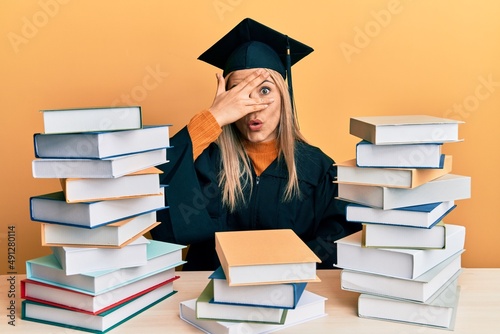 Young caucasian woman wearing graduation ceremony robe sitting on the table peeking in shock covering face and eyes with hand, looking through fingers with embarrassed expression.