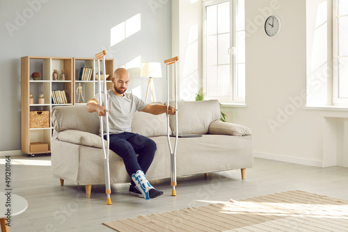 Canvastavla Young man with leg injury leans on crutches while trying to get up from sofa at home