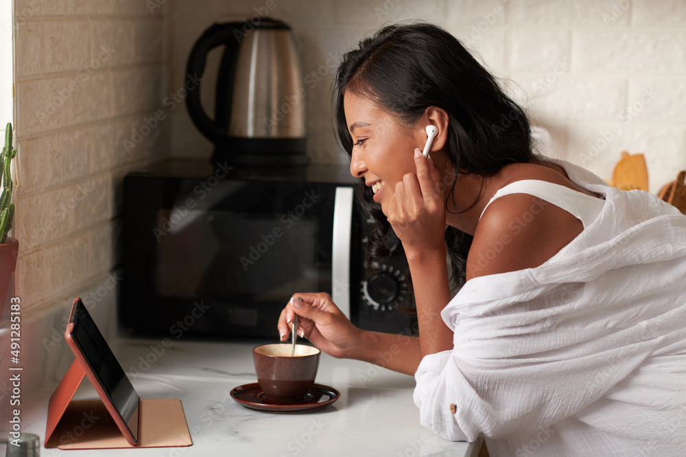 Happy young woman mixing sugar in her coffee when attending online briefing in the morning