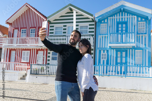Tourist couple taking a selfie hugging in front of the typical and well-known houses of Costa Nova in Portugal.