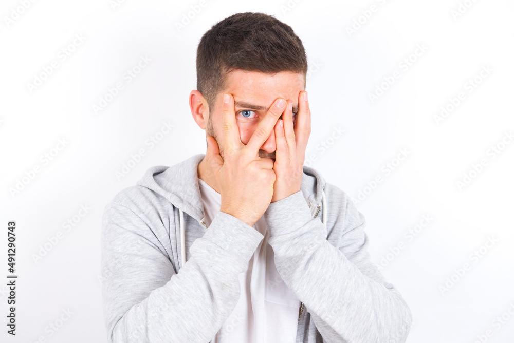 young caucasian man wearing casual clothes over white background covering face with hands and peering out with one eye between fingers. Scared from something or someone. young caucasian man wearing casual clothes over white background covering face with hands and peering out with one eye between fingers. Scared from something or someone.