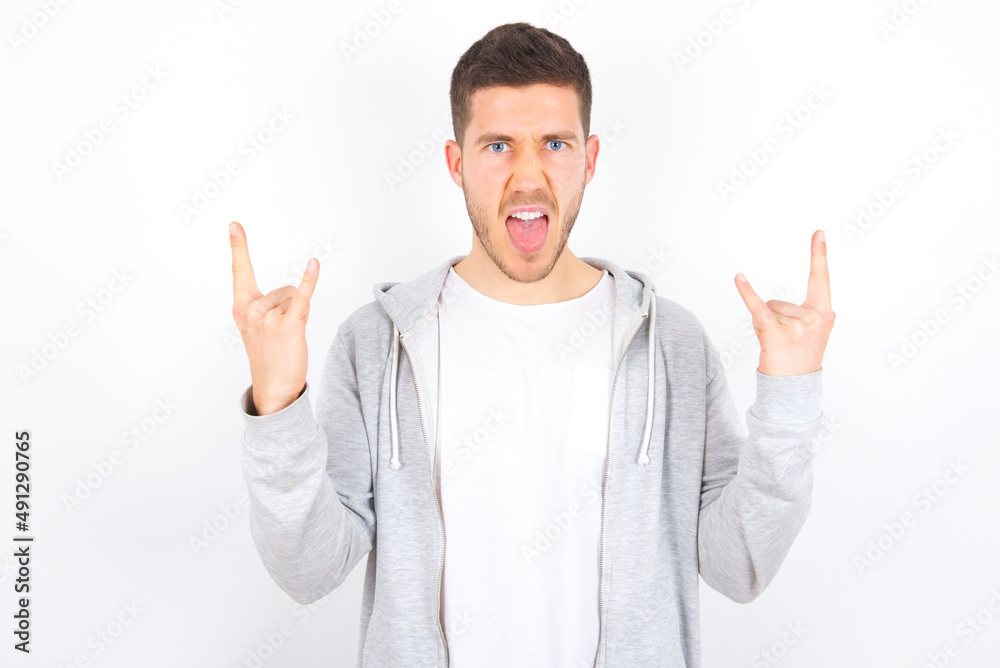Born to rock this world. Joyful young caucasian man wearing casual clothes over white background screaming out loud and showing with raised arms horns or rock gesture.