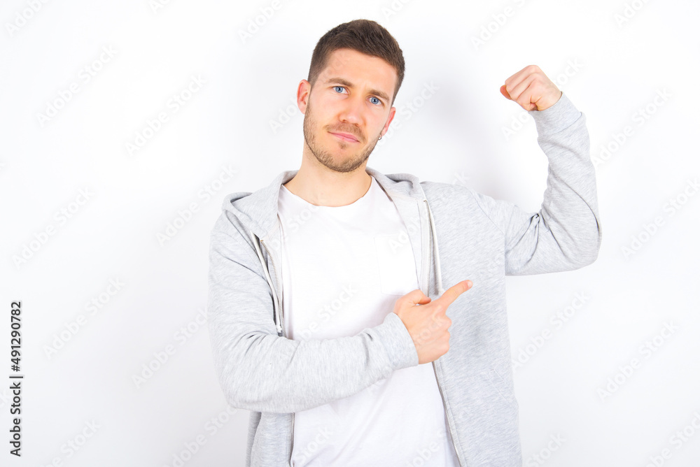 Smiling young caucasian man wearing casual clothes over white background raises hand to show muscles, feels confident in victory, strong and independent. Smiling young caucasian man wearing casual clothes over white background raises hand to show muscles, feels confident in victory, strong and independent.