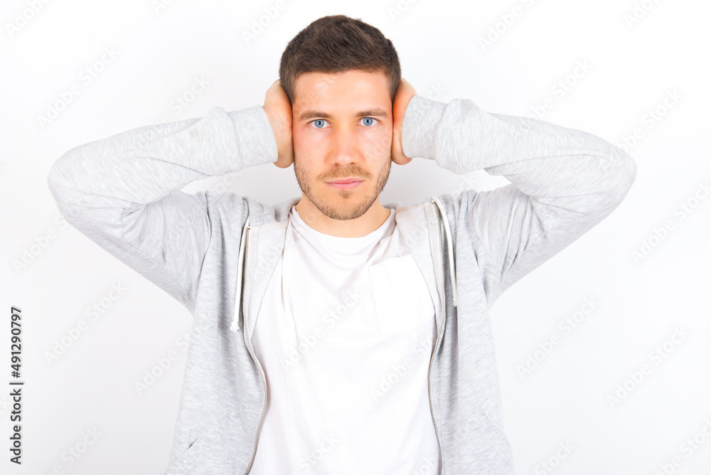 Frustrated young caucasian man wearing casual clothes over white background plugging ears with hands does not wanting to listen hard rock, noise or loud music. Frustrated young caucasian man wearing casual clothes over white background plugging ears with hands does not wanting to listen hard rock, noise or loud music.
