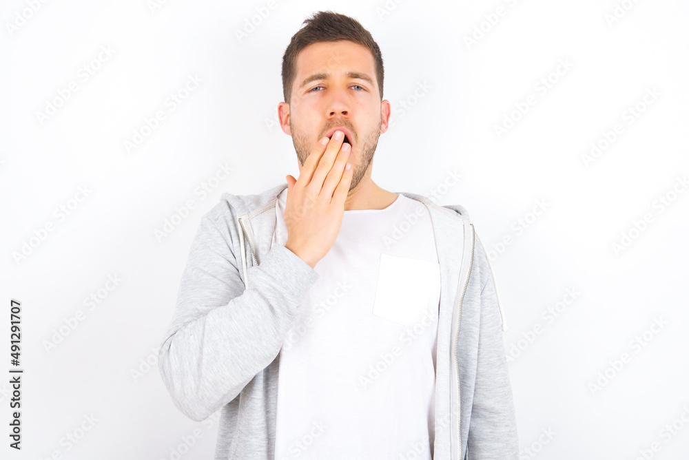 Sleepy young caucasian man wearing casual clothes over white background yawning with messy hair, feeling tired after sleepless night, yawning, covering mouth with palm. Sleepy young caucasian man wearing casual clothes over white background yawning with messy hair, feeling tired after sleepless night, yawning, covering mouth with palm.