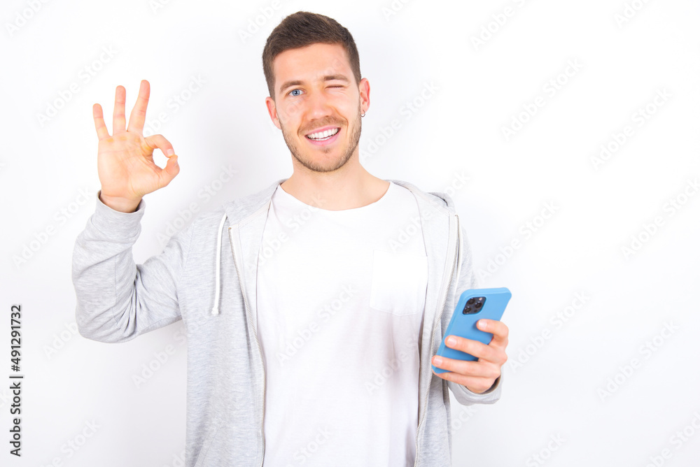 Happy young caucasian man wearing casual clothes over white background sending a message on his smartphone or taking a selfie and making ok sign with his hand. Happy young caucasian man wearing casual clothes over white background sending a message on his smartphone or taking a selfie and making ok sign with his hand.
