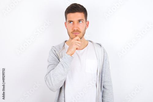 Thoughtful young caucasian man wearing casual clothes over white background holds chin and looks away pensively makes up great plan