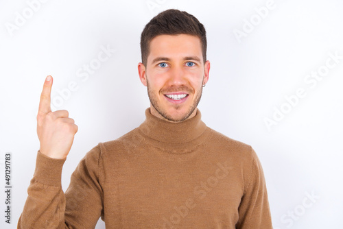 young caucasian man wearing knitted turtleneck over white background smiling and looking friendly, showing number one or first with hand forward, counting down