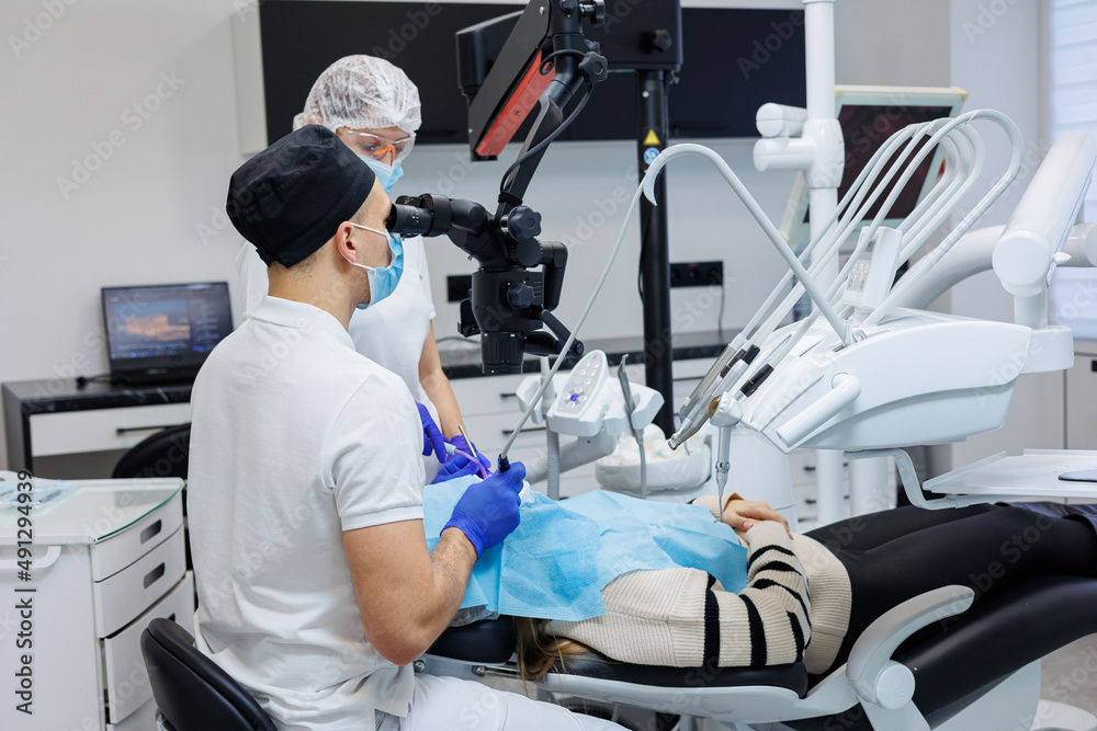 A successful dentist looks at the patient's teeth with a dental microscope and holds dental instruments near his mouth. The assistant helps the doctor. They wear white uniforms with masks and gloves. A successful dentist looks at the patient's teeth with a dental microscope and holds dental instruments near his mouth. The assistant helps the doctor. They wear white uniforms with masks and gloves.