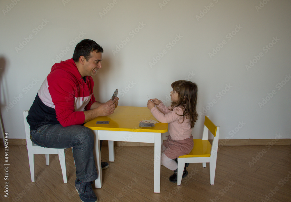 Happy father and daughter in the room while playing cards sitting on small children's chairs. Happy father and daughter in the room while playing cards sitting on small children's chairs.