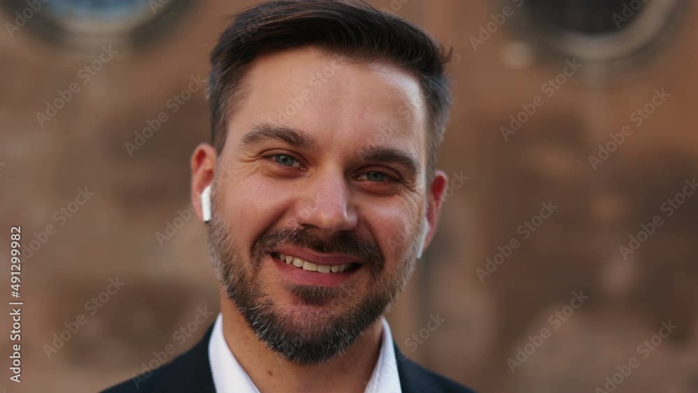Portrait view of the handsome man wearing earphones looking at the camera with pleasure smile while listening music at the street Portrait view of the handsome man wearing earphones looking at the camera with pleasure smile while listening music at the street