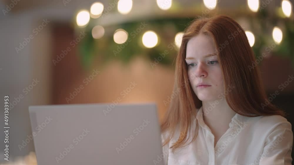 thoughtful brooding remote working red haired woman sitting infront of a laptop or notebook in casual outfit on her work desk in her modern airy bright living room home office with many windows thoughtful brooding remote working red haired woman sitting infront of a laptop or notebook in casual outfit on her work desk in her modern airy bright living room home office with many windows