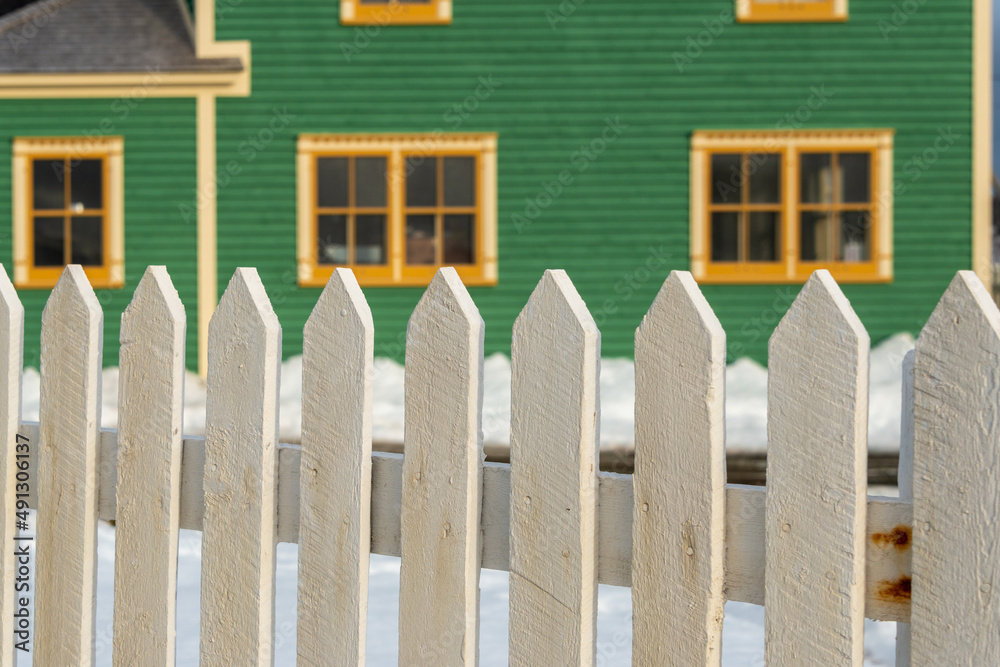 The exterior of a green cape cod clapboard horizontal wooden board style siding wall with two bright cream trim windows. There's snow on the ground and a white wooden picket fence in the foreground. The exterior of a green cape cod clapboard horizontal wooden board style siding wall with two bright cream trim windows. There's snow on the ground and a white wooden picket fence in the foreground.