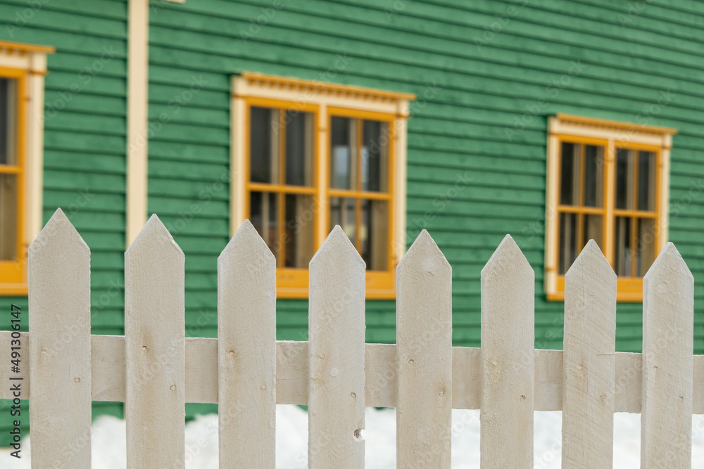 The exterior of a green cape cod clapboard horizontal wooden board style siding wall with two bright cream trim windows. There's snow on the ground and a white wooden picket fence in the foreground. 