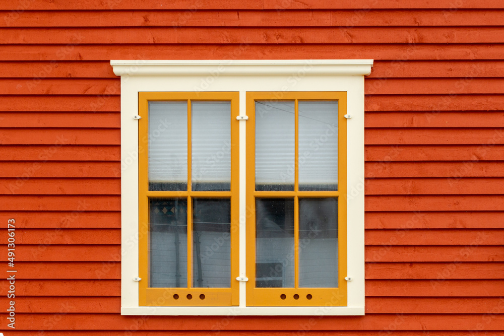 An exterior wall with orange colored horizontal cape cod clapboard siding and a double hung window. The closed vintage glass window has yellow and cream colored trim and molding with a window blind.