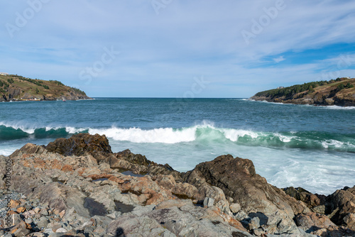 A seascape of a rocky beach with lots of sand and rocks on a bright sunny day. The blue sky, ocean, and horizon have a spray of mist from the rolling waves.  The shoreline is covered in beach rocks.