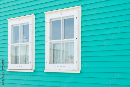 Two double hung windows on a green exterior wall of a vintage style building. The building has narrow horizontal clapboard siding. There s white wooden shutter and trim around the closed glass window.