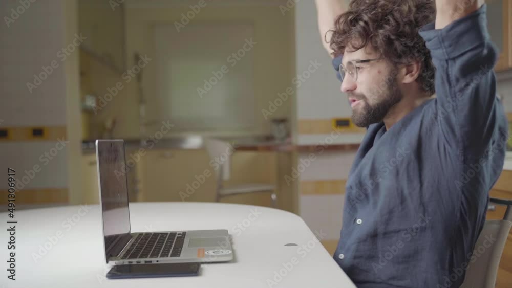 Exulting man with arms up working on laptop from home, sitting in yellow kitchen, caucasian Young adult model with beard and monitor glass.  Succeed, winning and cheerfulness concept