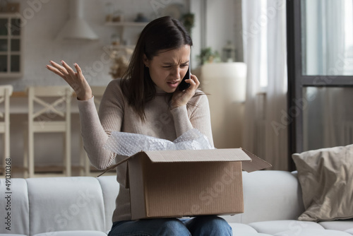 Woman sit on sofa open parcel box looks inside check purchased damaged items feels angry, complaining speaks on smartphone to customer support. Broken goods, bad services, dissatisfied client concept