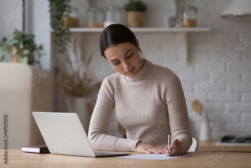 Tableau sur toile Young woman sit at table with laptop in kitchen holds pen, take notes on paper, writes letter