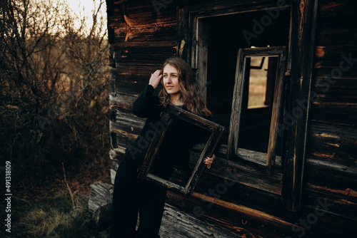 beautiful Ukrainian girl in black clothes near the old wooden house. The war in Ukraine. Portrait of a woman on a dark wooden background. Old abandoned wooden house. Old wooden window frame © Anhelina Tyshkovets