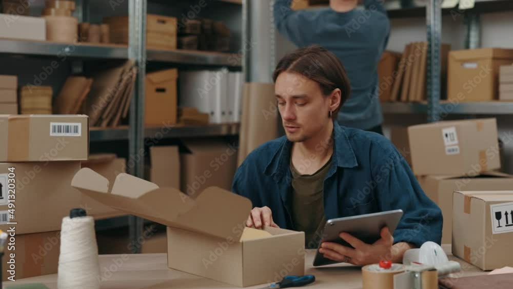 Stylish caucasian man sitting at the warehouse and looking at the tablet computer while typing parcel details on it. Post service and small business concept. Stylish caucasian man sitting at the warehouse and looking at the tablet computer while typing parcel details on it. Post service and small business concept.