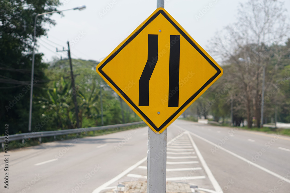 Yellow traffic square shaped left narrow lane sign at rural road Thailand to aware driver know Road narrows on left side ahead. Concept: Warning traffic sign for transportation. Yellow traffic square shaped left narrow lane sign at rural road Thailand to aware driver know Road narrows on left side ahead. Concept: Warning traffic sign for transportation.