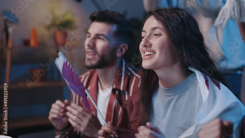 Close up of excited girl with flag rejoicing while watching sport on tv. Focus on woman with flags smiling while sitting on sofa with overjoyed friends nearby. Sport fans concept. photo
