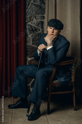 A young man with a cane is sitting in an armchair. English retro gangster of the 1920s dressed in a suit and flat cap in Peaky blinders style