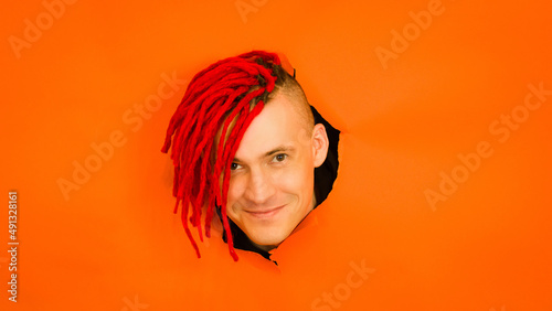Smiling man with dreadlocks looking at camera. Cheerful young male with dyed hair looking at camera in the hole on orange background in studio
