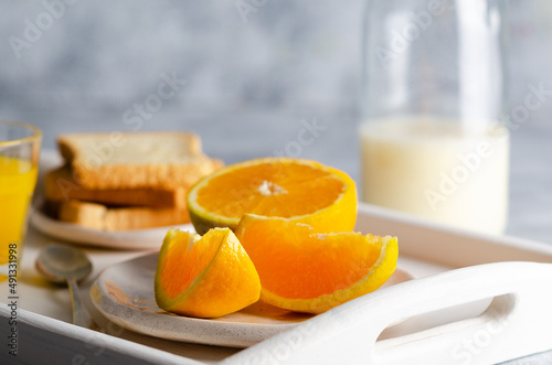 A bottle of milk  a glass of orange juice  and pieces of orange  in a white tray and grey background. 