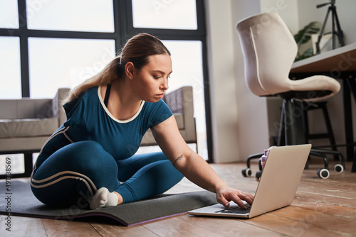 Tableau sur toile Woman with amputee limb sitting at the yoga mat and typing something at the lapt