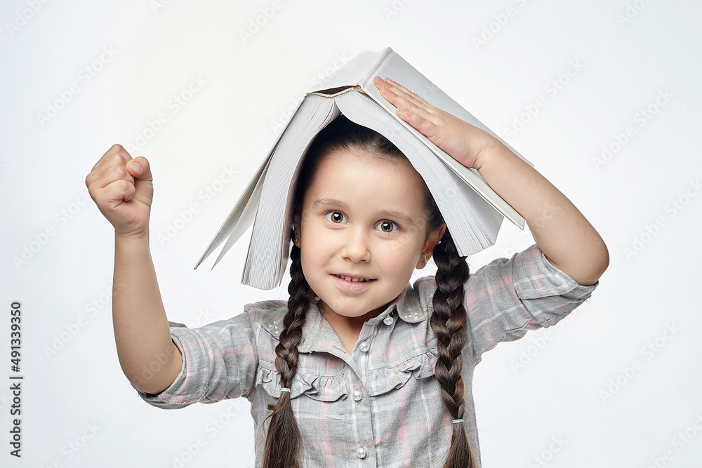 smiling little girl with braided pigtails shows her fist while holding a large book over her head smiling little girl with braided pigtails shows her fist while holding a large book over her head