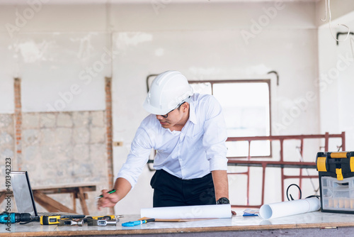 Civil construction engineer working with laptop at desk office with white yellow safety hard hat at office on construction site. Asian young man architecture project manager sitting at office on site