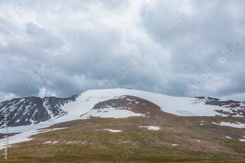 Dramatic landscape with large snowy mountain dome in sunlight under cloudy sky. Colorful scenery with sunlit high snow mountain in dome shape in center at changeable weather. Scenic mountains view.