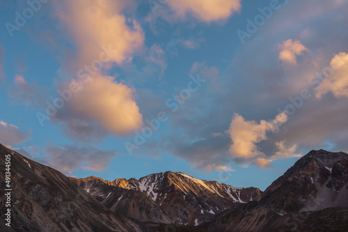 Scenic landscape with high snow mountain with sharp rocky pinnacle in golden sunlight under clouds of sunset color at changeable weather. Colorful view to large mountain top under orange clouds in sky