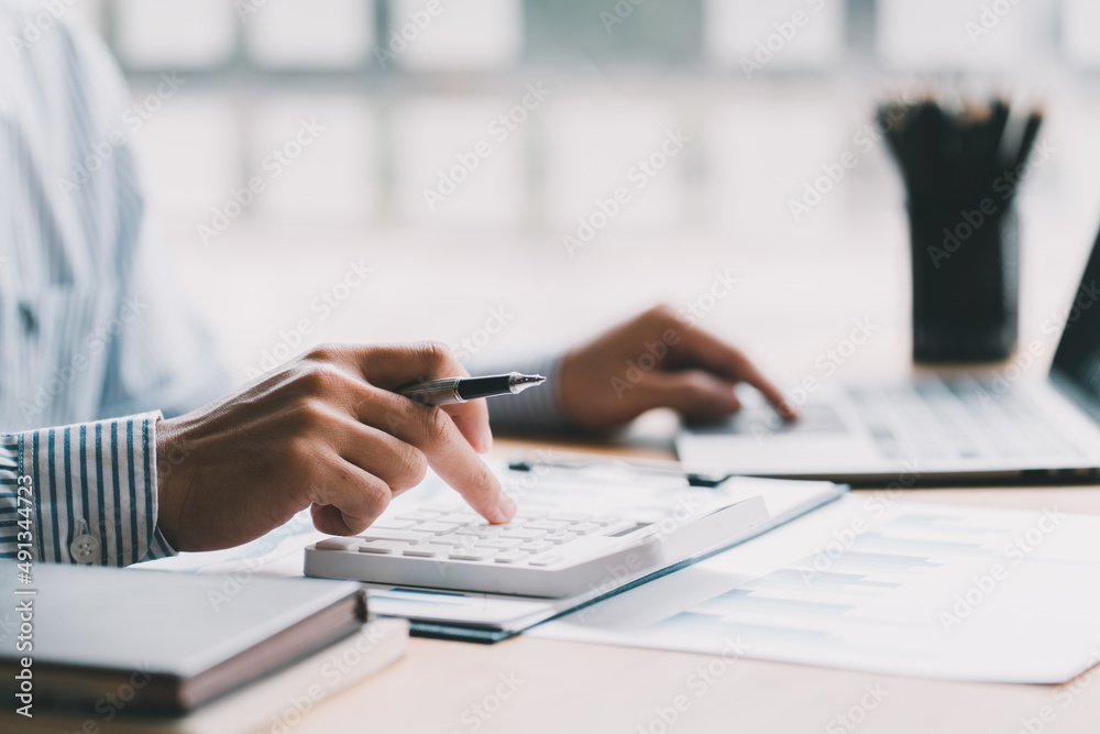 Close up of businessman or accountant hand holding pencil working on calculator to calculate financial data report, accountancy document and laptop computer at office, business concept