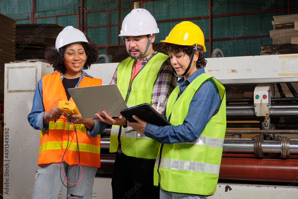 Safety uniform workers and industrial engineers with hardhat use laptop computer to check and control machines. Three professionals work in paper manufacturing factory, maintain production equipment. Safety uniform workers and industrial engineers with hardhat use laptop computer to check and control machines. Three professionals work in paper manufacturing factory, maintain production equipment.