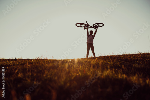Cyclist is holding his bicycle over himself on the background of red sunset. Biker with bicycle on the field during sunrise