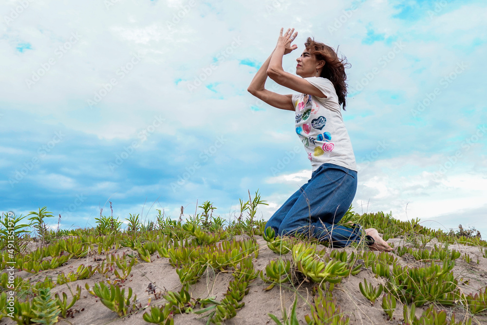 Adult latin woman smiling and doing yoga in the beach receiving the spring season in a praying pose and close eyes, feeling the fresh air. Freedom and joyful lifestyle concept. Adult latin woman smiling and doing yoga in the beach receiving the spring season in a praying pose and close eyes, feeling the fresh air. Freedom and joyful lifestyle concept.