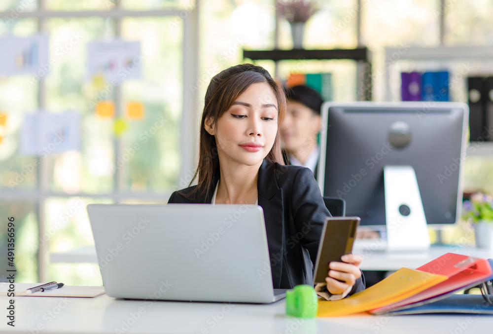Fototapeta premium Asian young female professional businesswoman secretary employee in formal suit sitting at working desk typing report via laptop notebook computer while businessman manager work behind in office