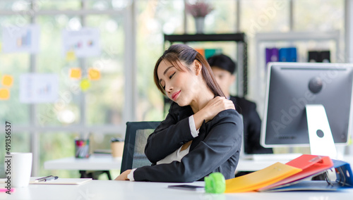 Asian young tired exhausted female professional businesswoman secretary in formal suit sitting relaxing at work desk holding hand massaging rubbing neck back and shoulder stretching muscle in company