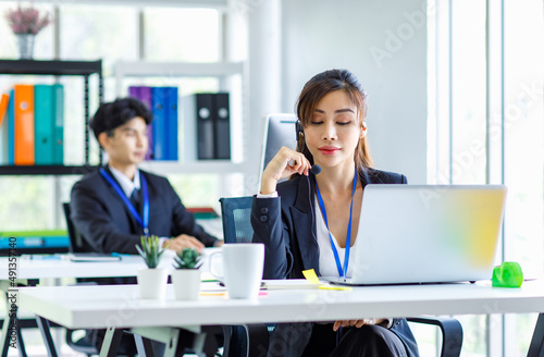 Asian young beautiful female businesswoman operator wearing headset with microphone working at helpdesk service with male colleague via laptop computer helping consulting with customers call center