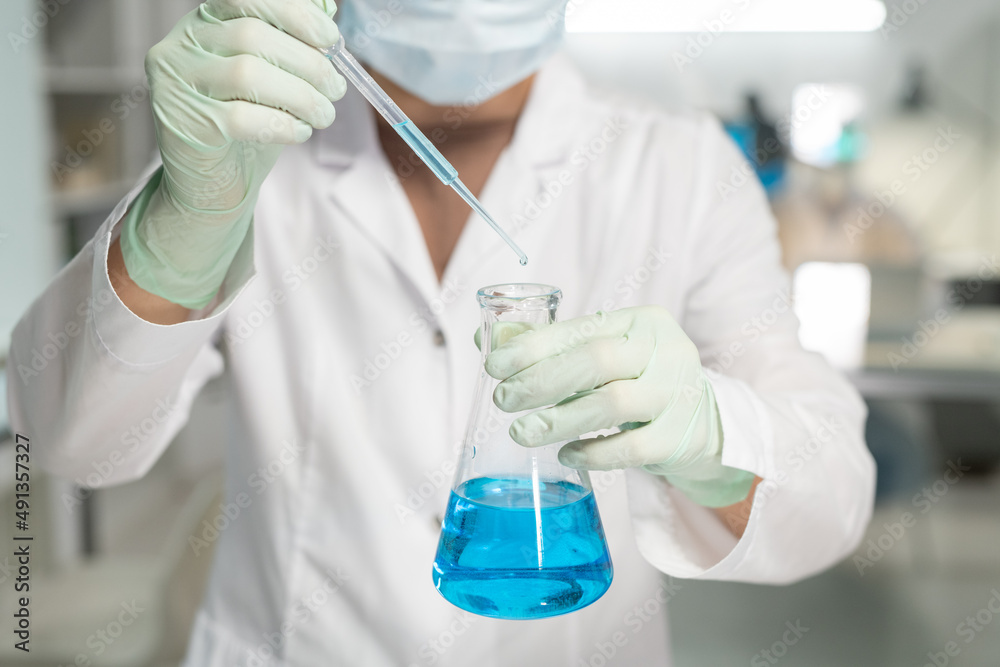 Contemporary scientist in surgical gloves, mask and lab coat adding drop of liquid into flask with blue chemical substance Contemporary scientist in surgical gloves, mask and lab coat adding drop of liquid into flask with blue chemical substance
