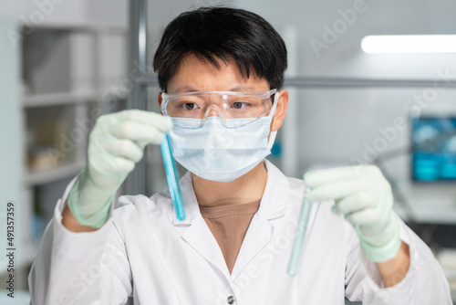 Asian female scientist in protective eyeglasses  mask and gloves comparing two samples of liquid substances