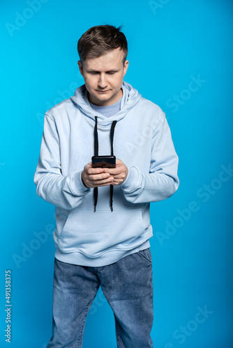 a guy in a blue sweatshirt and jeans is holding a phone. youth with gadget. blue background © Ольга Новицкая