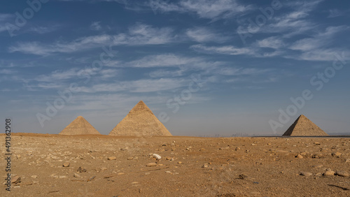 Three Great Pyramids Giza against blue sky and clouds. Stones are scattered on the sand of the desert. Silhouettes of modern buildings of Cairo are visible on the horizon. Egypt