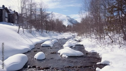 Amazing drone footage. Flying low over snow capped rocks in a creek in a mountainous region of Norway. Cabins are also present on the shore of the creek. Beautiful, sunny winter day with blue sky. photo