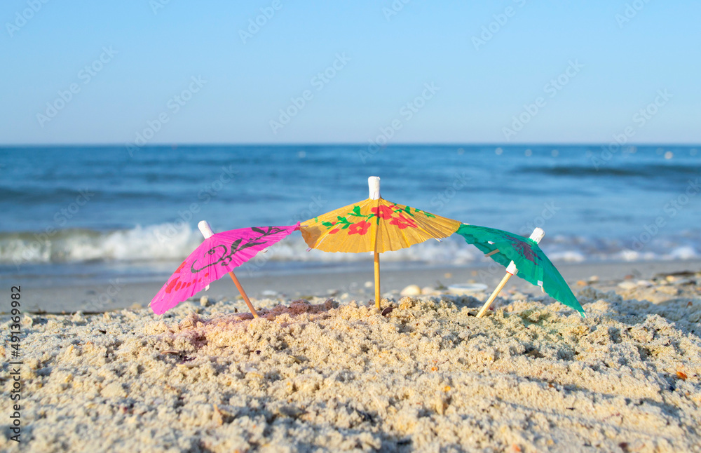 Three small paper cocktail umbrellas stand in sand on sandy beach close-up. Small paper umbrellas on sandy shore near sea and sea waves on sunny summer day. Leisure, Vacation, Travel, Tourism Concept